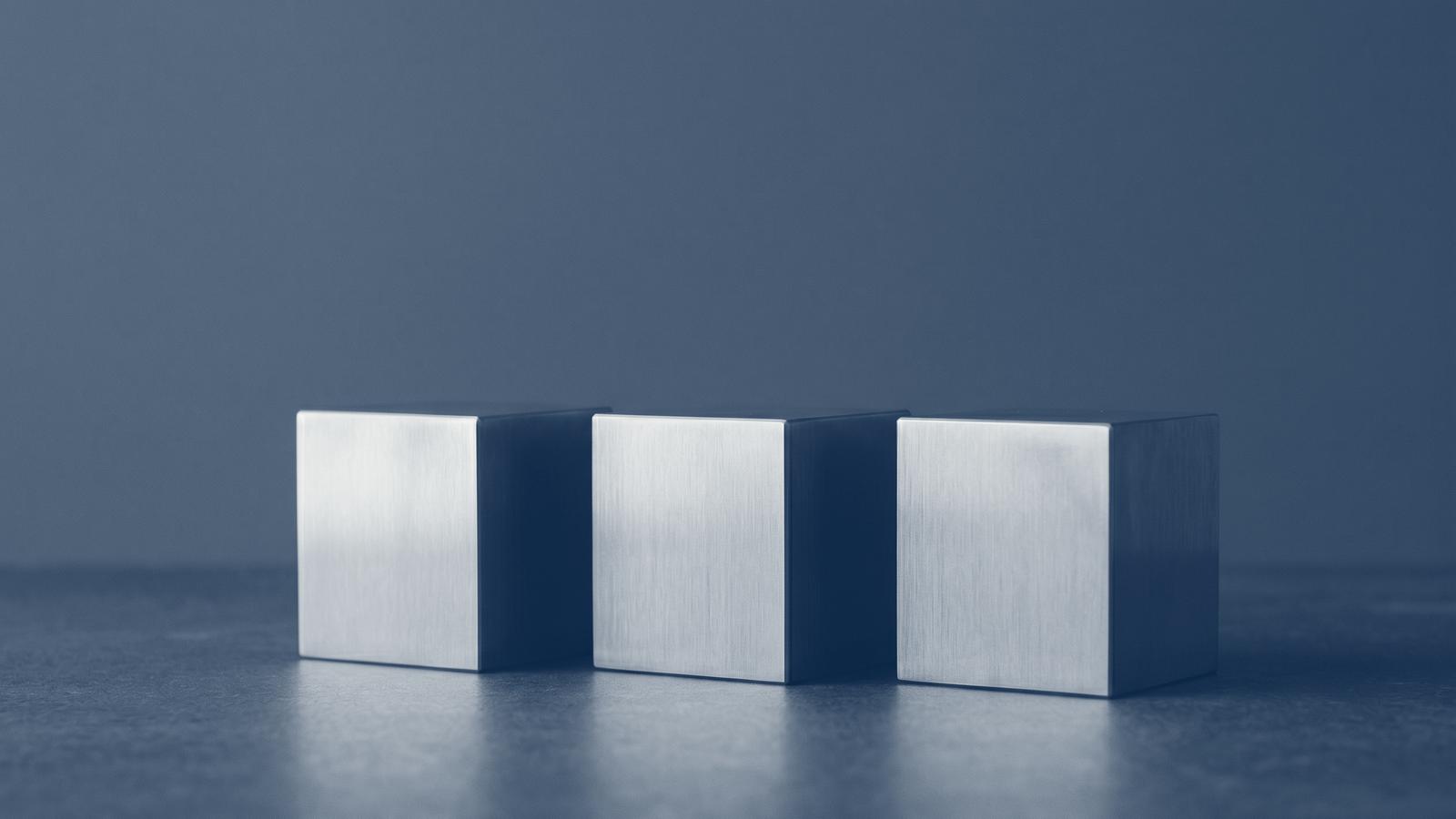 Three identical brushed-steel cubes in a row on a dark surface, toned navy: distributed, independent, equal.